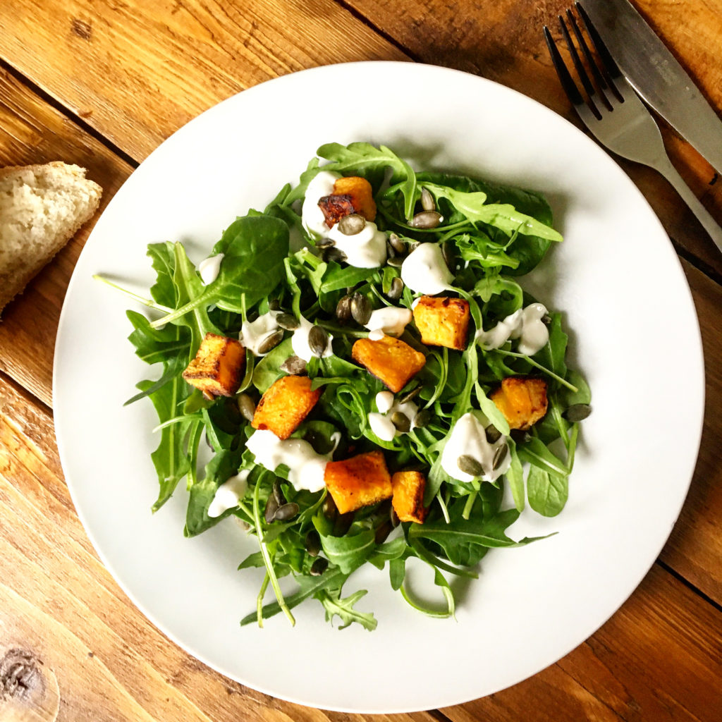 White plate of salad made from roasted butternut squash, spiniach, rocket, pumpkin seeds and a yoghurt dressing
