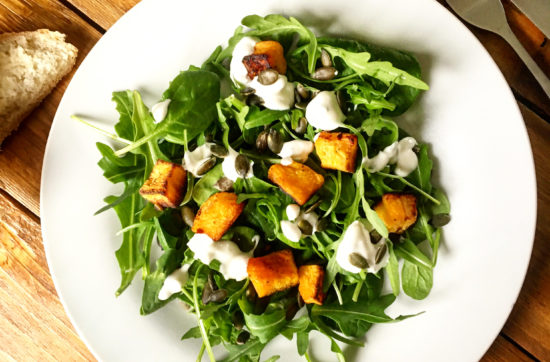 White plate of salad made from roasted butternut squash, spiniach, rocket, pumpkin seeds and a yoghurt dressing