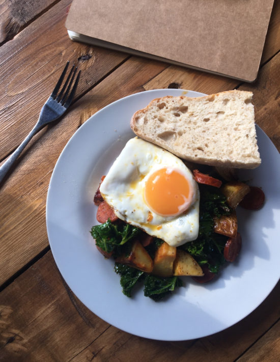 Chorizo, kale and potato hash topped with a fried egg and served with fresh baguette, served on a white plate on a wooden table