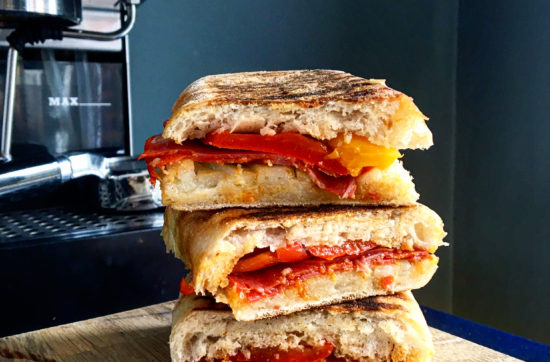 Stack of three pieces of chorizo, pepper and manchego panini ona wooden board in front of a Gaggia coffee machine