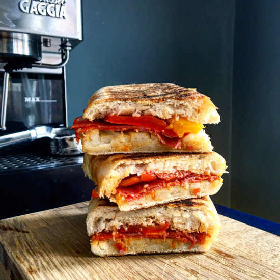 Stack of three pieces of chorizo, pepper and manchego panini ona wooden board in front of a Gaggia coffee machine