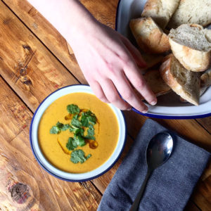 Bowl of carrot soup topped with corinader, on a wooden table and a hand reaching for fresh bread