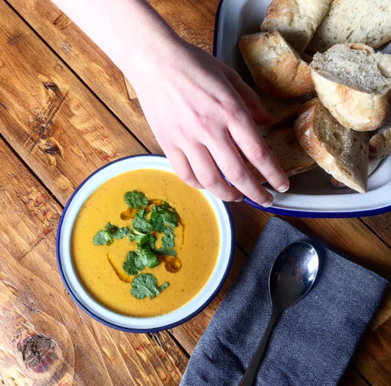 Bowl of carrot soup topped with corinader, on a wooden table and a hand reaching for fresh bread