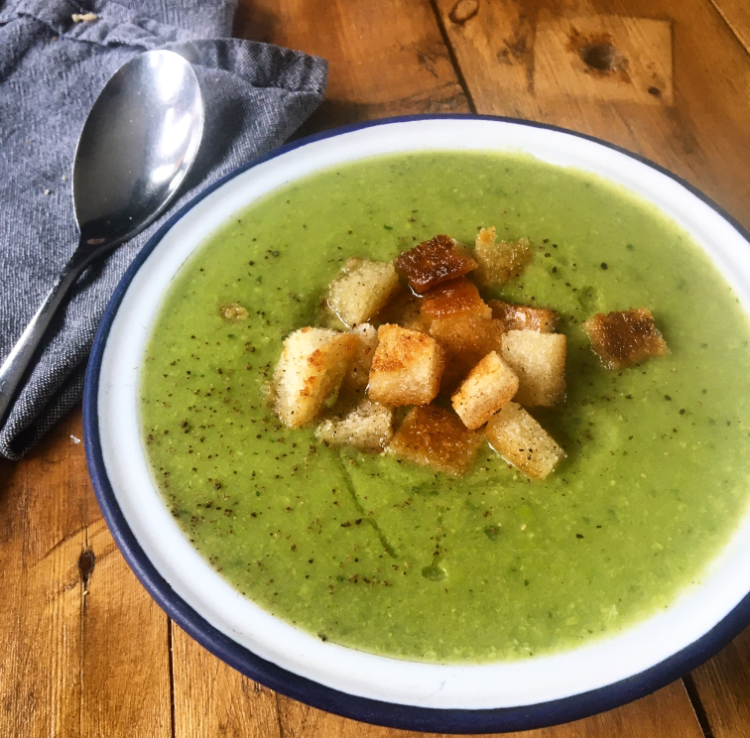 a bowl of minted pea soup with croutons, on a wooden table next to a spoon and napkin