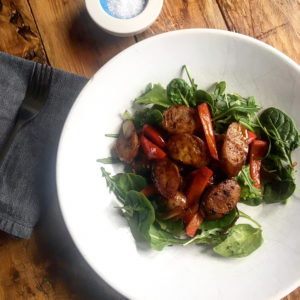 Bowl of mixed leaf salad topped with sliced sausages, red peppers, onions and balsamic dressing