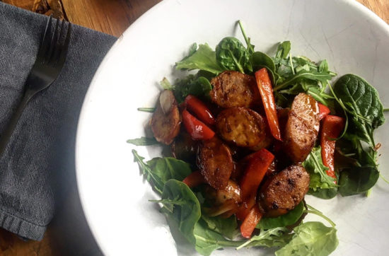 Bowl of mixed leaf salad topped with sliced sausages, red peppers, onions and balsamic dressing