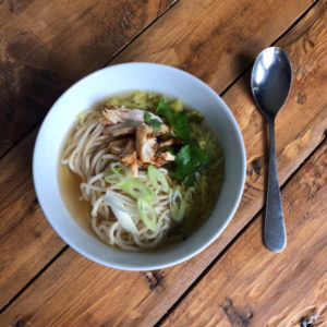 A bowl of Ramen with noodles and chicken, on a wooden table with a silver spoon