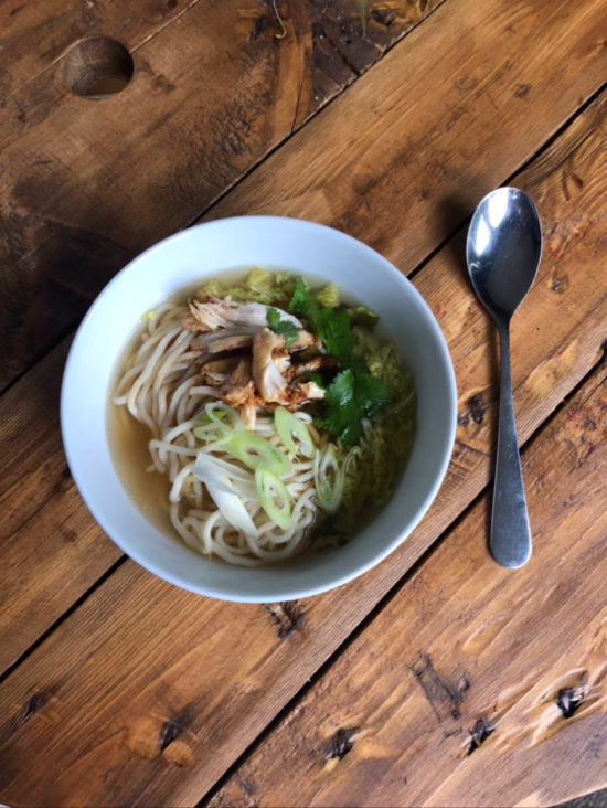 A bowl of Ramen with noodles and chicken, on a wooden table with a silver spoon