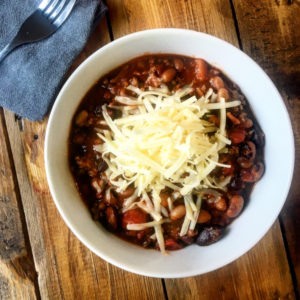 Bowl of beef and bean chilli topped with grated cheese, served in a white bowl on a wooden table, with a fork and napkin to the left
