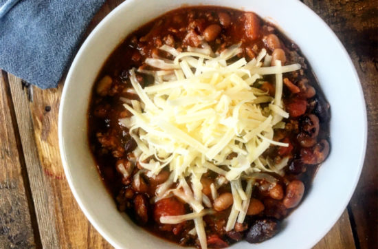 Bowl of beef and bean chilli topped with grated cheese, served in a white bowl on a wooden table, with a fork and napkin to the left