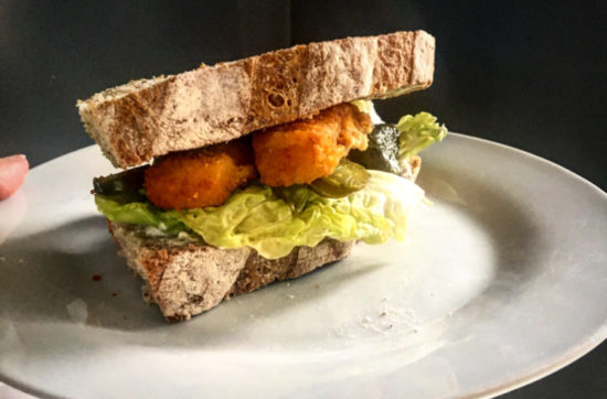 White plate serving a fish finger sandwich, with lettuce, gherkins, mayonnaise on crusty bread.