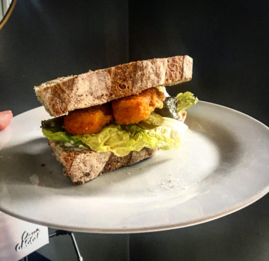 White plate serving a fish finger sandwich, with lettuce, gherkins, mayonnaise on crusty bread.