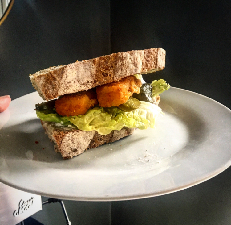 A white plate with a sandwich on top, made of crusty bread, lettuce, fish fingers, gherkins and mayonnaise.