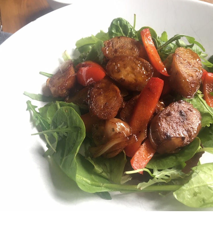 Mixed leaf salad topped with sliced sausages and balsamic dressing, served in a white bowl