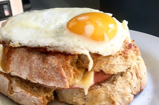 White plate topped with two layers of toasted white thickly-cut bread, with ham and cheese, topped with a runny fried egg