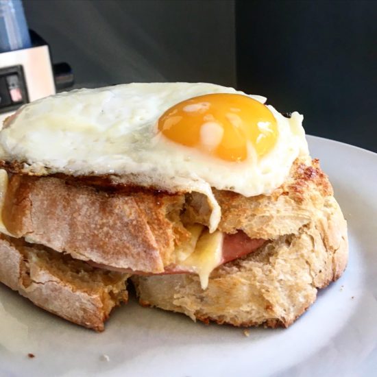 White plate topped with two layers of toasted white thickly-cut bread, with ham and cheese, topped with a runny fried egg