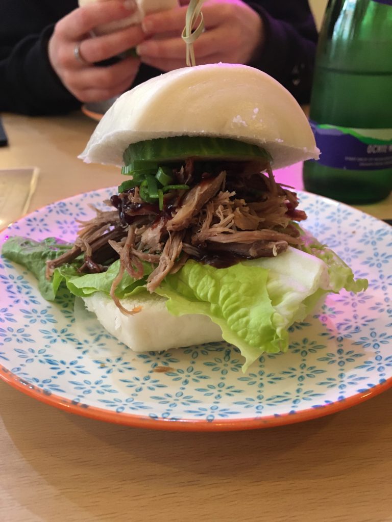 Photo of a bao bun on a plate filled with hoisin duck, cucumber and lettuce