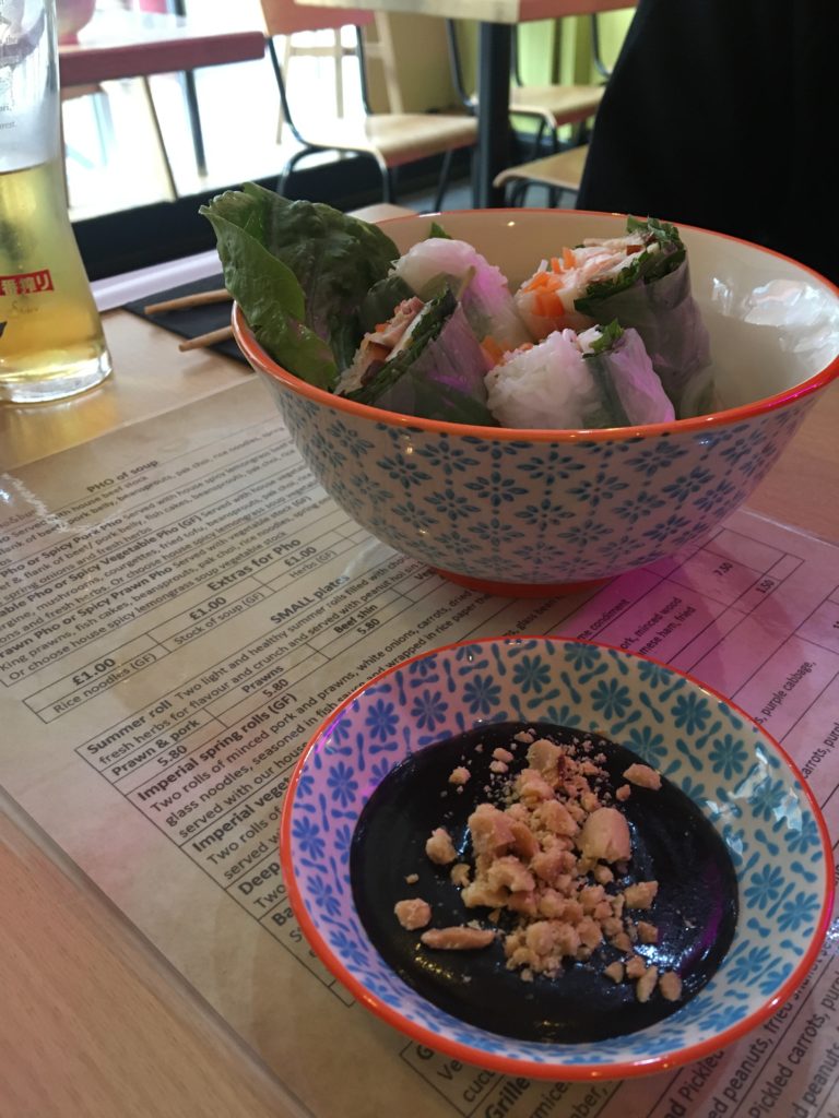 Photo of a table with two bowls. One serving Vietnamese summer rolls. The other, a dipping sauce topped with peanuts.