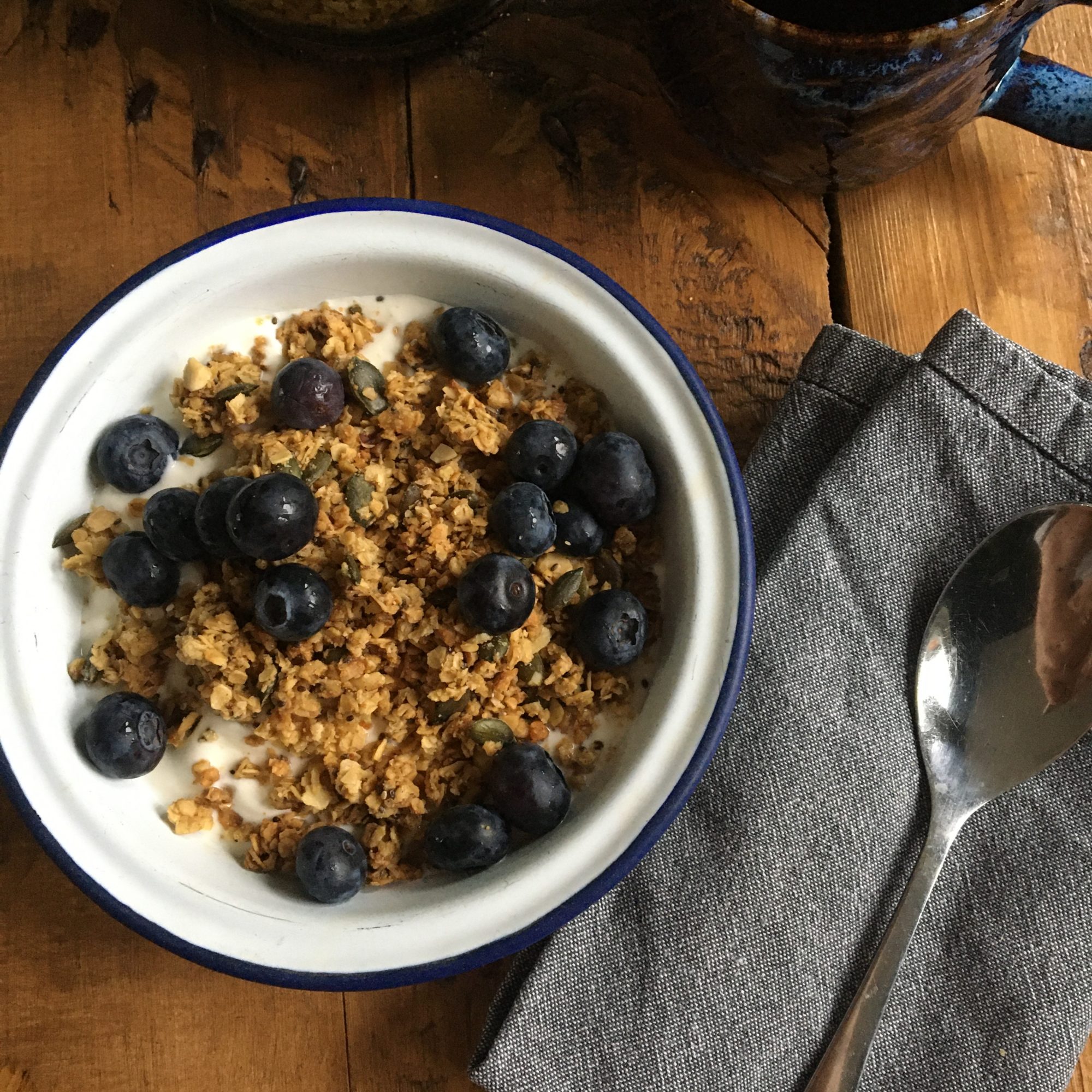 Photo shows an overhead shot of a blue and white enamel bowl full of granola, blueberries and yoghurt, with a blue napkin and silver spoon to the side