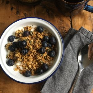 Photo shows an overhead shot of a blue and white enamel bowl full of granola, blueberries and yoghurt, with a blue napkin and silver spoon to the side